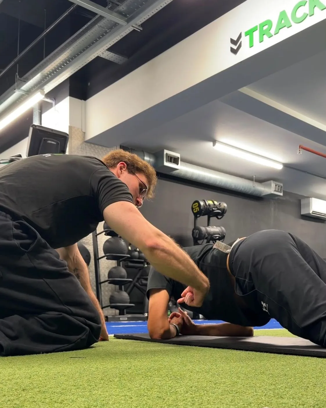 Manuel kneeling beside a client to correct their plank position on the gym floor